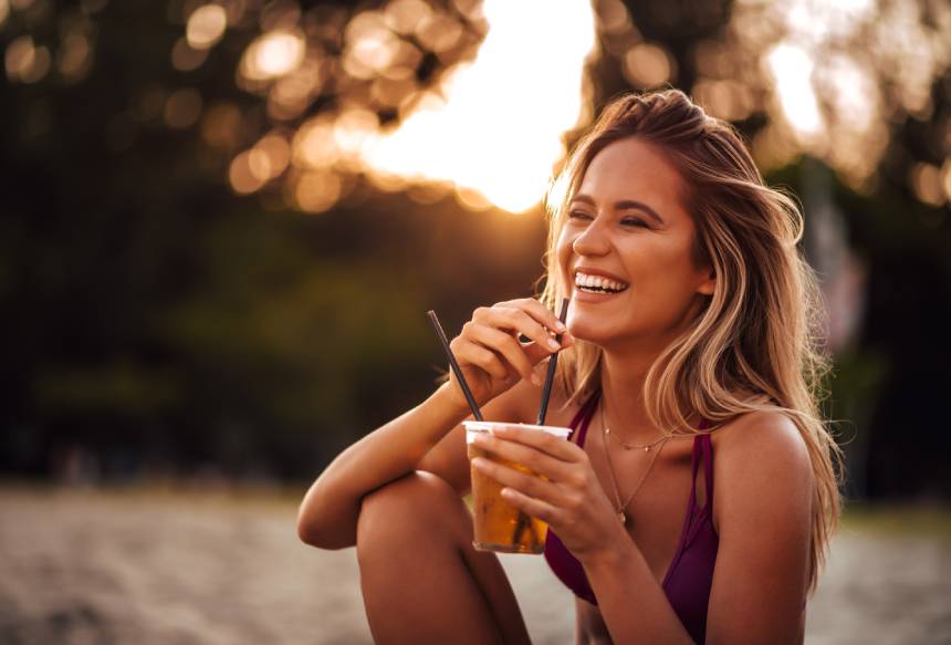 A beautiful, happy young blonde woman drinking on a beach during golden hour.